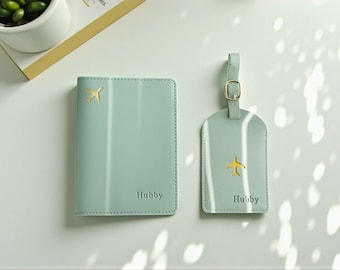 May include: A light green passport holder and luggage tag set. Each item features a gold airplane emblem and the word "Hubby". The passport holder is rectangular, and the luggage tag has a strap with a gold buckle.