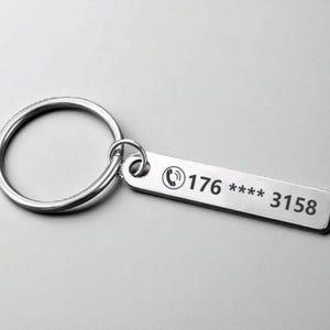 Könnte beinhalten: Ein silberfarbener Schlüsselanhänger mit einem rechteckigen Anhänger und einem großen kreisförmigen Ring. Der Anhänger hat ein Telefonsymbol und die Nummer "176 **** 3158" eingraviert. Der Schlüsselanhänger befindet sich auf einem hellgrauen Hintergrund.
