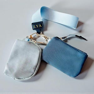 Puede incluir: Dos pequeñas bolsas con cremallera, una gris claro y otra azul, conectadas por cierres plateados a un cordón azul claro. Una pequeña etiqueta azul marino con las letras doradas "LYA" está unida al cordón.