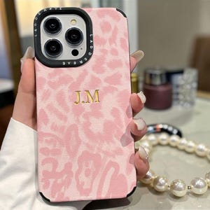 May include: Pink phone case with a leopard print design and the gold initials "J.M." The case has a black border around the camera and a black frame. The phone case is held in a hand.