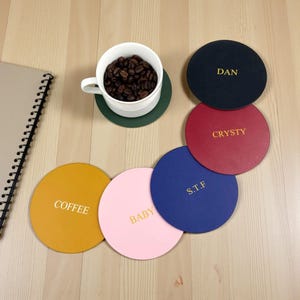 Könnte beinhalten: Eine Sammlung personalisierter runder Untersetzer in verschiedenen Farben, darunter Schwarz, Rot, Blau, Gelb und Rosa. Jeder Untersetzer hat goldfarbene Schriftzüge mit Namen oder Wörtern wie "DAN", "CRYSTY", "S.T.F", "COFFEE" und "BABY". Eine weiße Kaffeetasse mit Kaffeebohnen steht auf einem grünen Untersetzer.