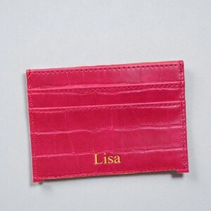 Puede incluir: Un tarjetero rosa brillante con un patr&oacute;n texturizado similar a la piel de cocodrilo. El tarjetero tiene m&uacute;ltiples ranuras y el nombre "Lisa" est&aacute; impreso en letras doradas. El art&iacute;culo es rectangular y parece estar hecho de cuero.