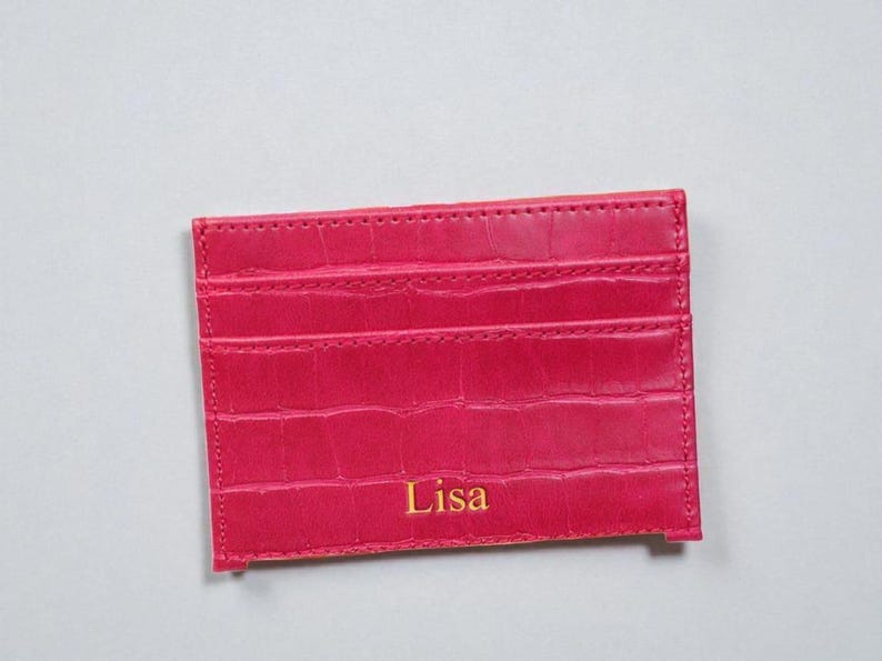 Puede incluir: Un tarjetero rosa brillante con un patr&oacute;n texturizado similar a la piel de cocodrilo. El tarjetero tiene m&uacute;ltiples ranuras y el nombre "Lisa" est&aacute; impreso en letras doradas. El art&iacute;culo es rectangular y parece estar hecho de cuero.