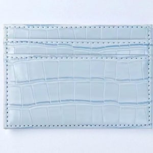 Puede incluir: Un tarjetero azul claro con textura de piel de cocodrilo. La cartera rectangular tiene m&uacute;ltiples ranuras para tarjetas y est&aacute; hecha de un material liso. Los bordes est&aacute;n cosidos con un hilo en contraste.