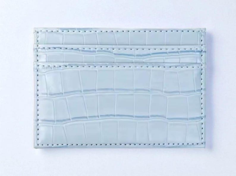 Puede incluir: Un tarjetero azul claro con textura de piel de cocodrilo. La cartera rectangular tiene m&uacute;ltiples ranuras para tarjetas y est&aacute; hecha de un material liso. Los bordes est&aacute;n cosidos con un hilo en contraste.
