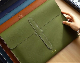 May include: A collection of leather document holders in various colors, including green, brown, blue, and black. The green holder in the foreground features a flap closure with a strap and the initials J.I.K.G. in gold. The image shows a hand touching the green holder.