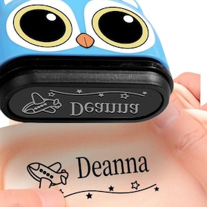 Könnte beinhalten: Ein blauer Stempel in Eulenform mit schwarzem Sockel stempelt den Namen "Deanna" und eine Flugzeuggrafik auf Stoff. Der Stempel trägt auch den Namen "De69UUS". Der Stempel wird von einer Person gehalten.