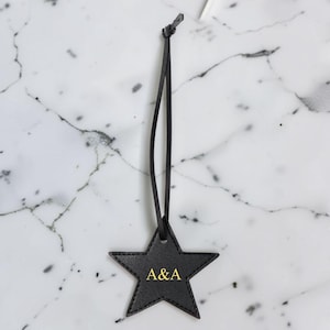 May include: A black star-shaped luggage tag with gold stitching and the initials "A&A" in gold lettering. The tag is suspended by a thin black cord. The background is a white marble surface.