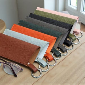 May include: A collection of leather eyeglass cases in various colors, including brown, orange, and gray, each with a metal keyring. One case displays the name "GILI". A pair of glasses rests in front of the cases.