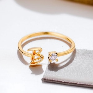 Puede incluir: Un anillo dorado con la letra "B" y una gema clara de corte cuadrado. El anillo está sobre una superficie blanca con una tela gris en el fondo. Una joya delicada.