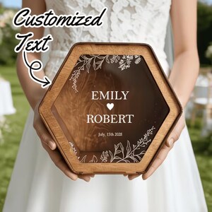 Puede incluir: Una caja hexagonal de madera con un panel frontal transparente que presenta arte lineal botánico blanco, un icono de corazón y el texto impreso "EMILY ROBERT July, 15th 2028". La caja tiene un acabado de veta de madera natural.