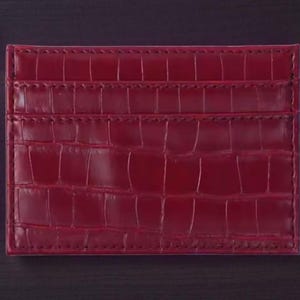 Puede incluir: Un tarjetero burdeos con textura de piel de cocodrilo. La cartera rectangular tiene m&uacute;ltiples ranuras para tarjetas y est&aacute; hecha de un material liso y brillante. Un bol&iacute;grafo con punta dorada es visible en el fondo.