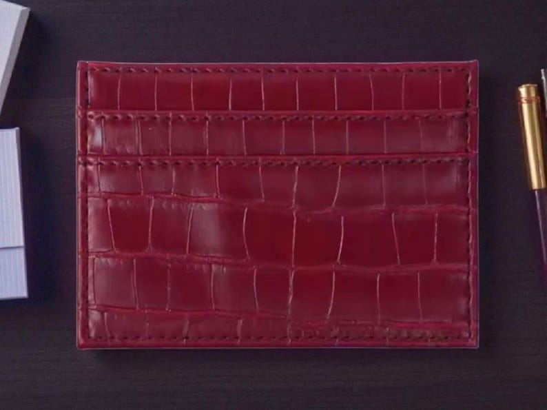 Puede incluir: Un tarjetero burdeos con textura de piel de cocodrilo. La cartera rectangular tiene m&uacute;ltiples ranuras para tarjetas y est&aacute; hecha de un material liso y brillante. Un bol&iacute;grafo con punta dorada es visible en el fondo.