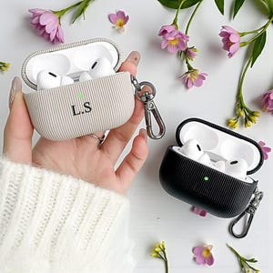 Könnte beinhalten: Zwei Schutzhüllen für kabellose Ohrhörer. Eine ist hellbeige mit den Initialen "L.S" und einem silbernen Verschluss. Die andere ist schwarz. Beide Hüllen haben eine gerippte Textur und sind geöffnet, wodurch die weißen Ohrhörer im Inneren sichtbar werden.