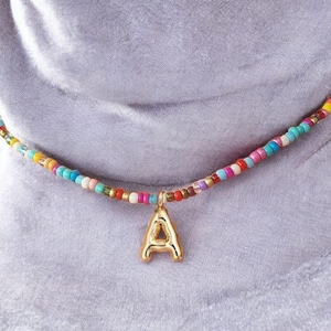 Op de afbeelding: Een kleurrijke kralenketting met een goudkleurige letter "A" hanger. De ketting heeft kleine, ronde kralen in verschillende kleuren, waaronder blauw, roze, geel en wit. De hanger is een gestileerde hoofdletter "A".