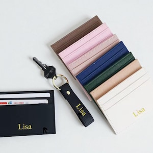 Könnte beinhalten: Eine Auswahl an Leder-Kartenhaltern in verschiedenen Farben, darunter Schwarz, Rosa, Blau und Beige. Jeder Kartenhalter ist mit dem Namen "Lisa" in goldfarbenen Buchstaben personalisiert. Ein schwarzer Schlüsselanhänger und ein Schlüssel sind ebenfalls vorhanden.