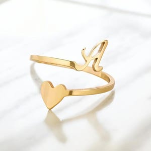 May include: A gold-coloured ring featuring a heart and the letter "A". The ring wraps around the finger, with the heart shape at one end and the letter "A" at the other. The ring is on a white surface.