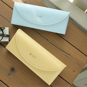 以下が含まれることがあります： 「Lily」と刻印された水色のケースと「RAY」と刻印された黄色のケースの2つのパーソナライズされたメガネケース。黒縁のメガネが白いブロックの上に置かれています。ケースは木製の表面にあります。