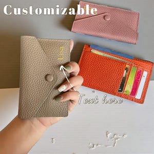 May include: A collection of leather cardholders in various colors. One is held in a hand, featuring a button closure and gold initials. The image includes a pink and orange cardholder, with the word "Customizable" and "Text here" in the background.