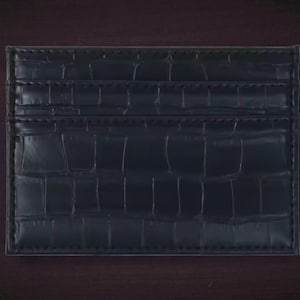 Puede incluir: Un tarjetero negro con textura de piel de cocodrilo. El tarjetero tiene m&uacute;ltiples ranuras para tarjetas y un dise&ntilde;o elegante y minimalista. El tarjetero est&aacute; hecho de un material duradero.
