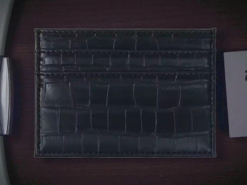Puede incluir: Un tarjetero negro con textura de piel de cocodrilo. El tarjetero tiene m&uacute;ltiples ranuras para tarjetas y un dise&ntilde;o elegante y minimalista. El tarjetero est&aacute; hecho de un material duradero.