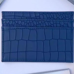 Puede incluir: Un tarjetero azul marino con textura de piel de cocodrilo. El tarjetero tiene tres ranuras y es rectangular. Las costuras son azul marino. El tarjetero est&aacute; sobre una superficie blanca.