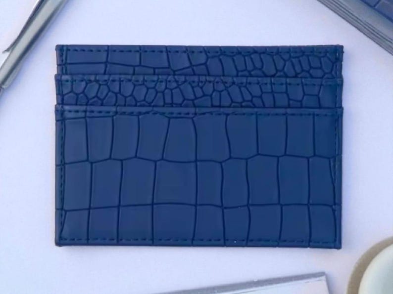 Puede incluir: Un tarjetero azul marino con textura de piel de cocodrilo. El tarjetero tiene tres ranuras y es rectangular. Las costuras son azul marino. El tarjetero est&aacute; sobre una superficie blanca.