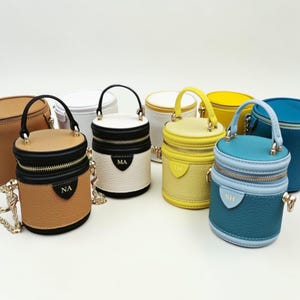 May include: A collection of small, cylindrical handbags in various colors, including tan, white, yellow, and blue. Each bag features a top handle, a zipper closure, and a small tag with initials. Some have chain straps.