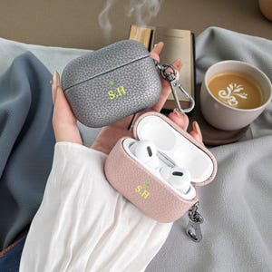 May include: Two personalized AirPod cases, one gray and one pink, with the initials "S.H." in green. The pink case is open, revealing white AirPods. Both cases have a textured leather appearance and a silver keychain clip.