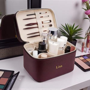 Può includere: Un beauty case in pelle bordeaux con interno color crema, aperto per rivelare pennelli per il trucco e vari prodotti cosmetici. Il beauty case è personalizzato con il nome "Lisa" in lettere dorate. Altri trucchi sono sul tavolo.