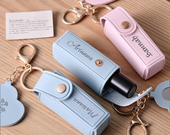 May include: A collection of personalized lipstick holders in pastel colors. The holders are light blue and pink, with gold-colored hardware and the names "Mariana", "Arianna", and "Sannah" engraved on them. Each holder is attached to a keychain.