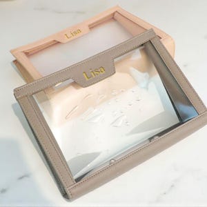 May include: Two clear cosmetic bags with the name "Lisa" in gold lettering. One bag is taupe with a clear front, and the other is light pink. The bags are rectangular with a clear front and a leather-like trim.