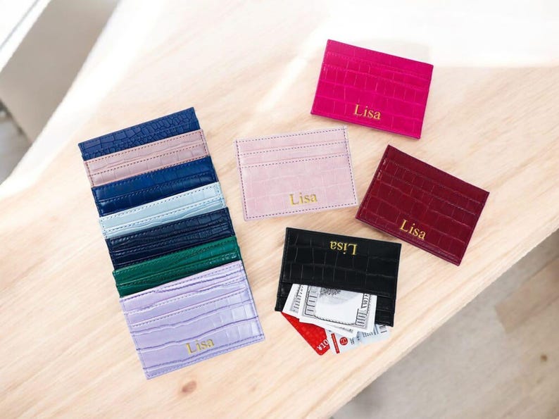 Puede incluir: Varios tarjeteros de cuero en colores como rosa, rojo, negro y azul. Cada tarjetero presenta un patr&oacute;n de cocodrilo texturizado y el nombre "Lisa" en letras doradas. Algunos tarjeteros est&aacute;n apilados, mientras que otros se muestran individualmente.