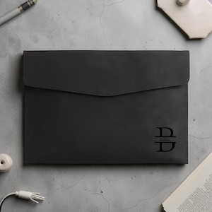 May include: A dark gray leather document holder with a flap closure. The holder features a monogram with the letter "D" and the name "Babbs" below it. The holder is on a gray surface with a pen and other objects.