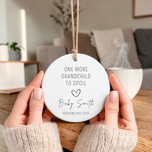 Op de afbeelding: Een witte keramische schijf met de tekst "ONE MORE GRANDCHILD TO SPOIL" en "Baby Smith ARRIVING MAY 2026" in zwart schrift. Een kleine hartafbeelding staat onder de tekst. De schijf is opgehangen aan een natuurlijk touwkoord.