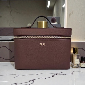 May include: A brown makeup case with a gold zipper and handle. The initials "G.G." are displayed on the front. Two perfume bottles are visible to the right of the case.