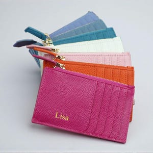 May include: A collection of colorful leather cardholders, each with a zipper closure and multiple card slots. The cardholders are in shades of pink, orange, white, teal, and blue. The front-most cardholder is a vibrant pink with the name "Lisa" in gold lettering.