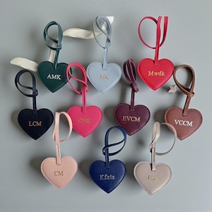 Può includere: Una collezione di etichette in pelle a forma di cuore in vari colori, ciascuna con un passante e un cinturino. Le etichette sono in rilievo con le iniziali in lettere dorate. I colori includono verde, blu, rosso, rosa e marrone.