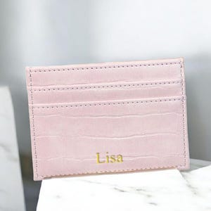 Puede incluir: Un tarjetero rosa claro con textura de cocodrilo. El tarjetero tiene tres ranuras para tarjetas y el nombre "Lisa" en letras doradas. El tarjetero es de cuero y es un accesorio elegante.