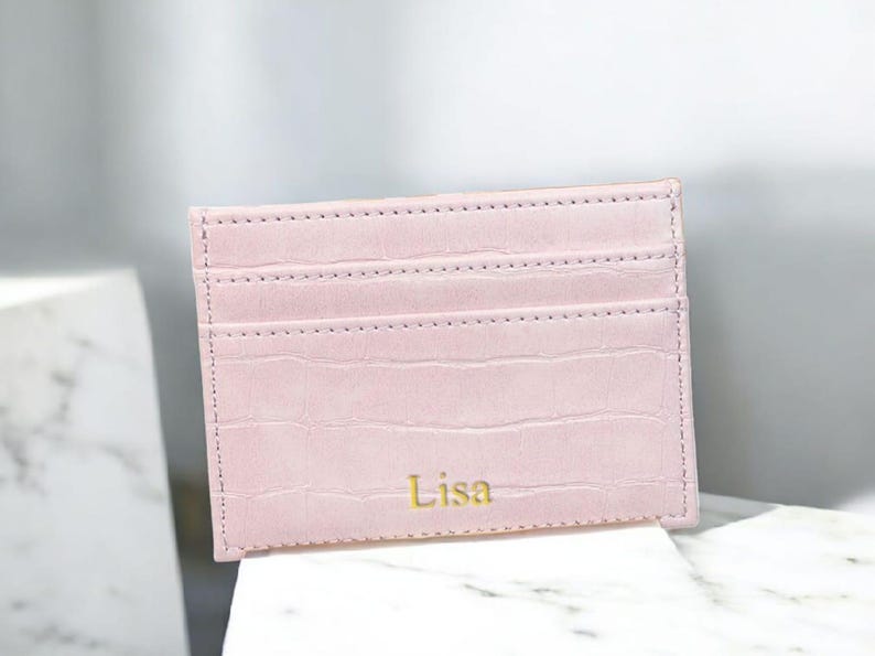 Puede incluir: Un tarjetero rosa claro con textura de cocodrilo. El tarjetero tiene tres ranuras para tarjetas y el nombre "Lisa" en letras doradas. El tarjetero es de cuero y es un accesorio elegante.