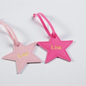May include: Two star-shaped luggage tags in shades of pink. Each tag has a loop for attachment and the name "Lisa" in gold lettering. The tags are made of a soft material, likely for personalising luggage or gifts.