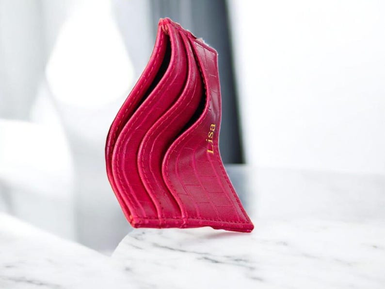Puede incluir: Un tarjetero fucsia con un patr&oacute;n texturizado similar al de un cocodrilo. El tarjetero tiene m&uacute;ltiples ranuras y el nombre "Lisa" est&aacute; impreso en dorado. El dise&ntilde;o es elegante y moderno, perfecto para llevar tarjetas.
