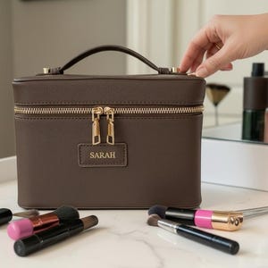 Op de afbeelding: Een bruine make-upkoffer met een gouden rits en handvat, gepersonaliseerd met de naam "SARAH". Make-upborstels en compacts zijn eromheen gerangschikt op een wit oppervlak. De koffer is ongeveer 20 cm breed.