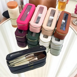 Può includere: Una collezione di astucci colorati per pennelli da trucco in vari colori, tra cui rosa, verde e marrone. Ogni astuccio ha una finestra trasparente e una chiusura con cerniera. Un astuccio è aperto, mostrando i pennelli da trucco. Gli astucci sono disposti su una superficie riflettente.