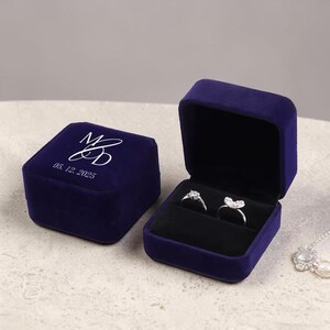 May include: Two open and closed navy blue velvet ring boxes. The closed box has the initials "MD" and the date "05. 12. 2025" in white. The open box displays two rings on a black interior.