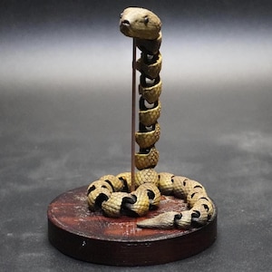 May include: A detailed sculpture of a snake with a coiled body and upright head. The snake is made of a light brown material with darker accents, and is mounted on a round, dark brown base. The snake's body is segmented.