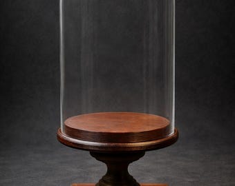 May include: A clear glass cloche with a rounded top, displayed on a dark wood base and pedestal. The base is square, and the pedestal has a decorative, turned design. The cloche is empty, allowing for display of an item.