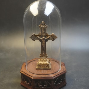 May include: A bronze crucifix displayed under a glass dome, presented on an octagonal wooden base. The cross has detailed carvings and depicts Jesus. The base is decorated with gothic-style carvings. The glass dome is transparent.