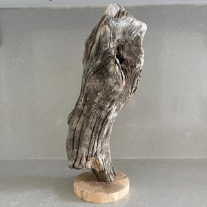 May include: A piece of weathered, grey driftwood with a textured, gnarled surface and a natural hole near the top, mounted vertically on a smooth, circular, light-colored wooden base. The wood shows deep grooves and organic, irregular contours.