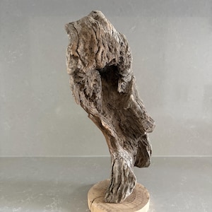 May include: A piece of weathered, textured driftwood sculpture mounted vertically on a circular, light-colored wooden base. The wood features deep grooves, natural cracks, and a gray-brown color, showcasing an organic, irregular, and hollowed-out form.